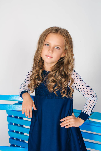 Cute girl with  blond curly hair in school fashion clothes with blue chair on a white background - Φωτογραφία, εικόνα