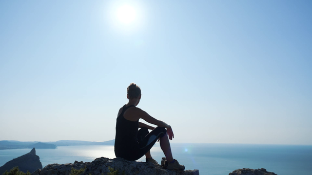 Joven mujer inspirada sentada en la cima de una montaña sobre el mar contra el hermoso cielo azul. Silueta de una mujer excursionista feliz sentada en la cumbre y disfrutando de una vista fantástica desde una altura
. - Metraje, vídeo