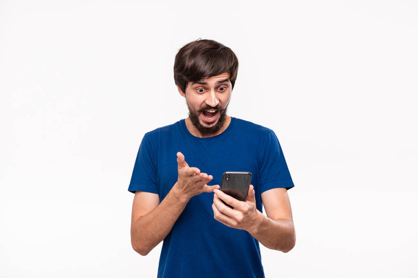 Όμορφος Μπρουνέ άνθρωπος με ένα μπλε πουκάμισο με γένια και μουστάκι έκπληκτος κοιτάζοντας την οθόνη ενός smartphone που στέκεται απομονωμένη σε λευκό φόντο. Συγκίνηση και χειρονομία σοκ. - Φωτογραφία, εικόνα