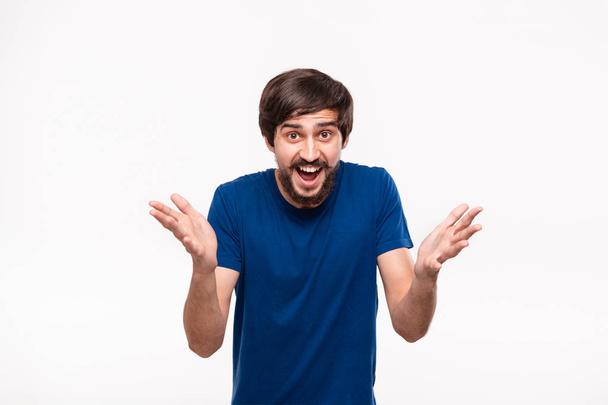 Good looking brunet man in a blue shirt with beard and mostaches shrugging his shoulders being surprised standing isolated over white background. Emotion and gesture of shock. - Foto, Bild