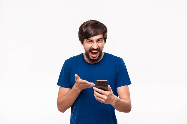Bello l'uomo brunetta in camicia blu con la barba e baffi scioccato guardando lo schermo di uno smartphone in piedi isolato su sfondo bianco. Emozione e gesto di sorpresa
. - Foto, immagini