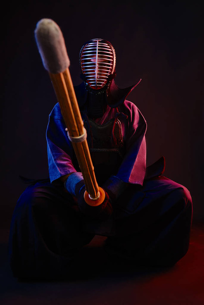 Close up. Kendo fighter wearing in an armor, traditional kimono, helmet, sitting, practicing martial art with shinai bamboo sword, black background. - Foto, imagen