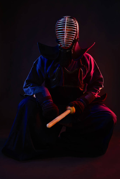 Close up. Kendo fighter wearing in an armor, traditional kimono, helmet, sitting, practicing martial art with shinai bamboo sword, black background. - Photo, Image