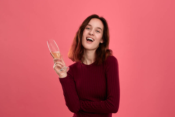 Ενθουσιασμένος όμορφη μελαχρινή κορίτσι κρατώντας κρασί της αφρώδης σαμπάνιας στέκεται απομονωμένη σε ένα σκούρο ροζ φόντο και χαμογελά στην κάμερα. Έννοια του εορτασμού - Φωτογραφία, εικόνα