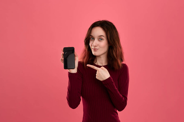 カメラを見て暗いピンクの背景に立っている間、指で携帯電話の画面を指で指で指で興奮かわいいブルネットの女の子. - 写真・画像
