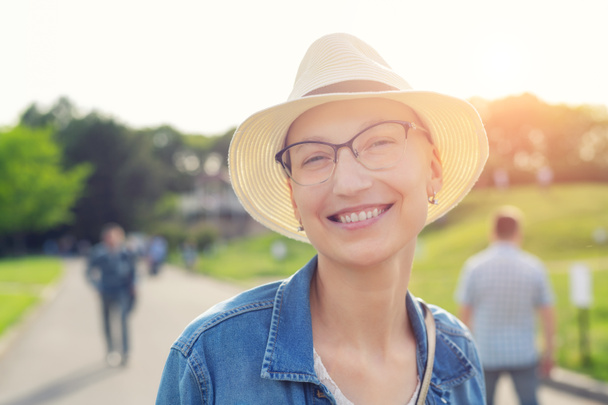 Szczęśliwa młoda, łysa kobieta w kapeluszu i luźnych ubraniach, ciesząca się życiem po przeżyciu raka piersi. Portret pięknej bezwłosej dziewczyny uśmiechniętej podczas spaceru po parku miejskim po leczeniu choroby - Zdjęcie, obraz