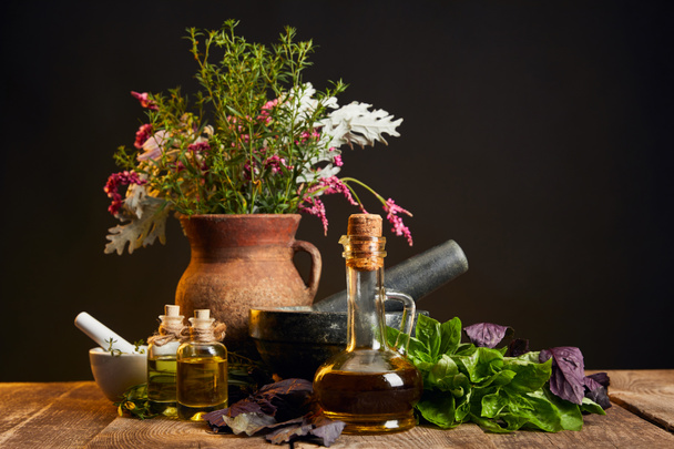 clay vase with fresh herbs and flowers near mortar and pestle and bottles on wooden table isolated on black - Photo, Image
