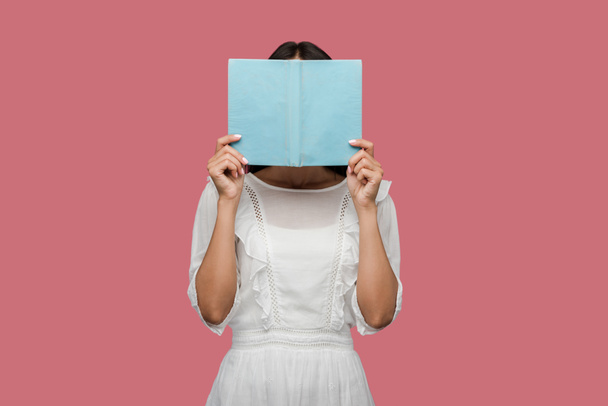 young woman in dress covering face with blue book isolated on pink  - Foto, Bild
