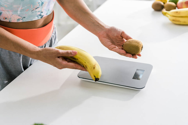 vista cortada de mulher segurando banana e kiwi perto de escalas de alimentos
 - Foto, Imagem