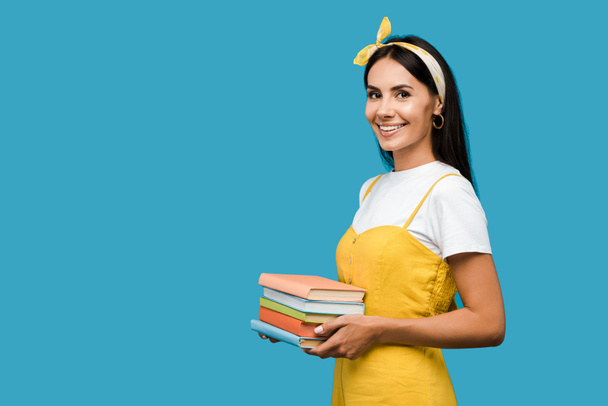 happy woman in headband holding books and looking at camera isolated on blue  - Fotoğraf, Görsel