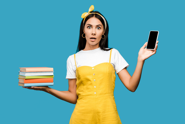 shocked woman holding books and smartphone with blank screen isolated on blue  - Foto, Imagen