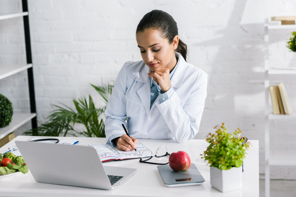 attractive woman in white coat writing in notebook near laptop and vegetables  - Photo, Image