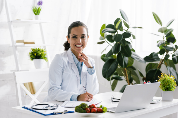 happy nutritionist in white coat near vegetables and laptop  - Photo, Image