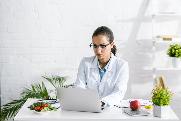attractive nutritionist in glasses using laptop near vegetables and plants  - Photo, Image