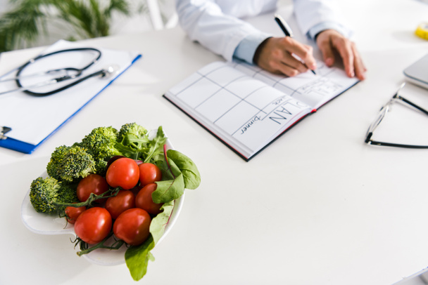 zugeschnittene Ansicht des Ernährungswissenschaftlers schreibt in Notizbuch mit Speiseplan in der Nähe von Gemüse  - Foto, Bild