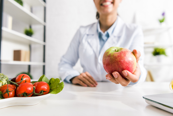 cropped view of nutritionist holding tasty apple near vegetables  - Photo, Image