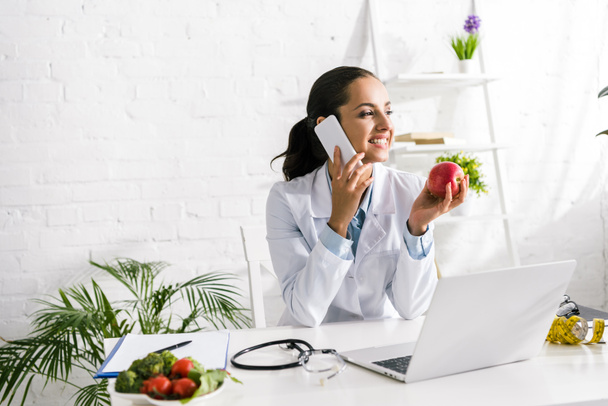 onnellinen ravitsemusterapeutti puhuu älypuhelimella läppärin ja omenan lähellä
  - Valokuva, kuva