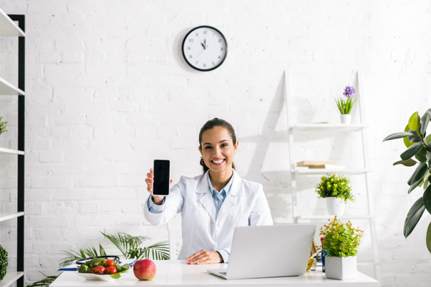 happy nutritionist holding smartphone with blank screen near laptop  - Photo, Image