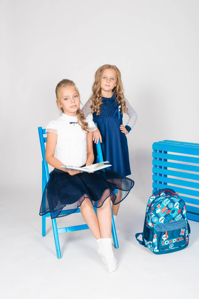 Two cute schoolgirls in a beautiful trendy school uniform with a backpack, books and notebooks with blue chair on a white background - Fotografie, Obrázek