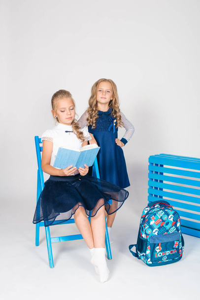 Two cute schoolgirls in a beautiful trendy school uniform with a backpack, books and notebooks with blue chair on a white background - Fotoğraf, Görsel