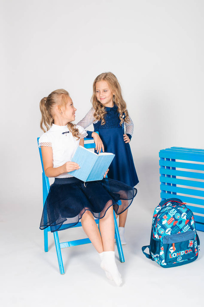 Two cute schoolgirls in a beautiful trendy school uniform with a backpack, books and notebooks with blue chair on a white background - Φωτογραφία, εικόνα