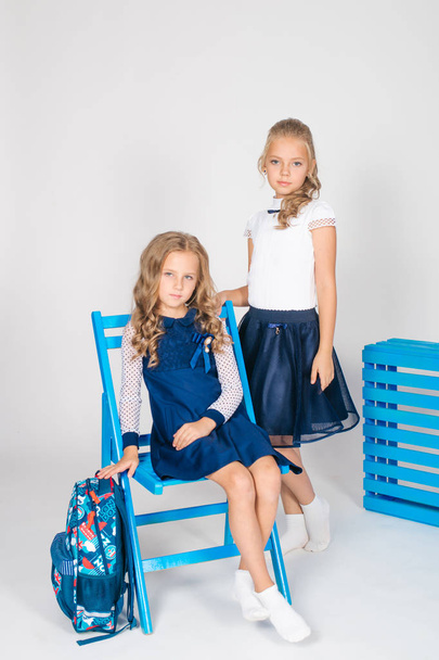Two cute schoolgirls in a beautiful trendy school uniform with a backpack, books and notebooks with blue chair on a white background - Фото, зображення