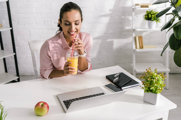 onnellinen nainen tilalla muovi kuppi appelsiinimehua lähellä kannettavaa tietokonetta ja omena
  - Valokuva, kuva