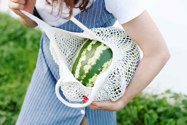 Cropped view of woman in denim dress posing with watermelon in knitted bag - Foto, Bild