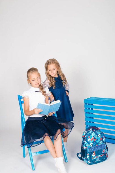 Two cute schoolgirls in a beautiful trendy school uniform with a backpack, books and notebooks with blue chair on a white background - Fotografie, Obrázek