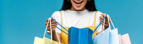 Panoramaaufnahme einer glücklichen jungen Frau mit bunten Einkaufstüten auf blauem Grund  - Foto, Bild