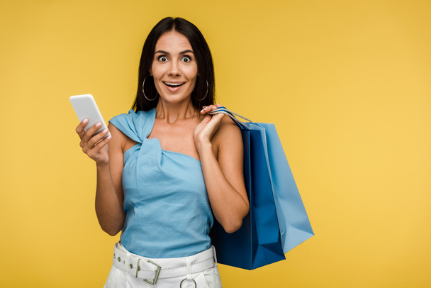 excited young woman looking at camera and holding smartphone with shopping bags isolated on orange - Fotografie, Obrázek