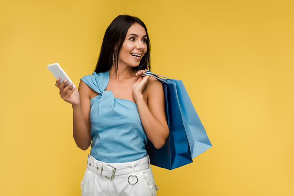 excited woman holding smartphone with shopping bags isolated on orange  - Foto, Imagem