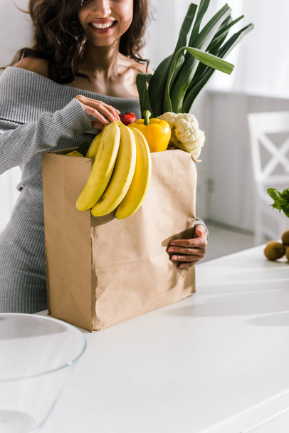cropped view of happy girl holding paper bag with groceries  - Фото, изображение