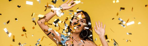panoramic shot of cheerful young woman gesturing near sparkling confetti on orange  - Photo, Image