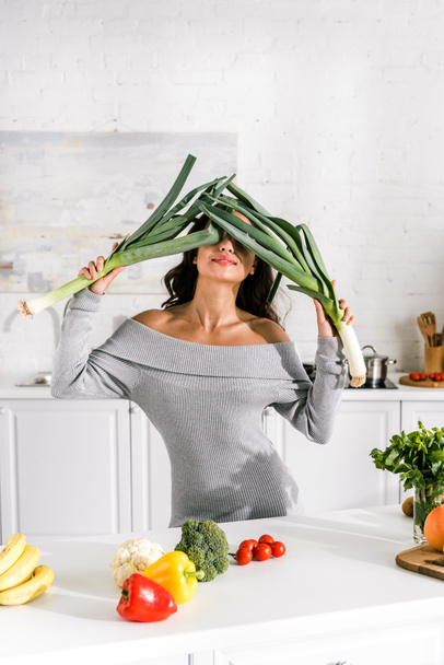 Atrakcyjna dziewczyna trzyma por w pobliżu twarzy i smaczne warzywa na stole  - Zdjęcie, obraz