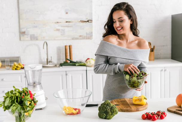 cheerful young woman holding greenery and looking at bowl with salad  - Photo, Image