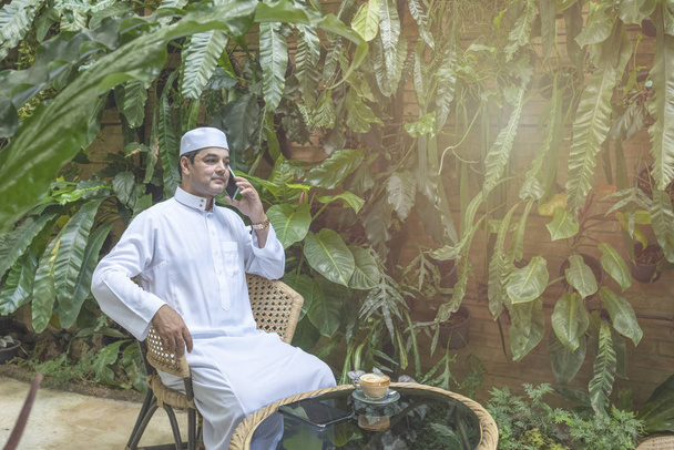 スマートケータイを使用してパキスタンのビジネスアジアの男性は、コーヒーショップに座ってイスラム教徒のドレスを着用深刻な顔で話す、フリーランスのビジネスコンセプト. - 写真・画像