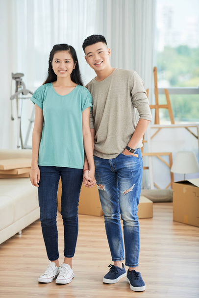 Portré fiatal ázsiai pár állva alkalmi ruházati kéz kezében, és mosolyogva a kamera otthon - Fotó, kép