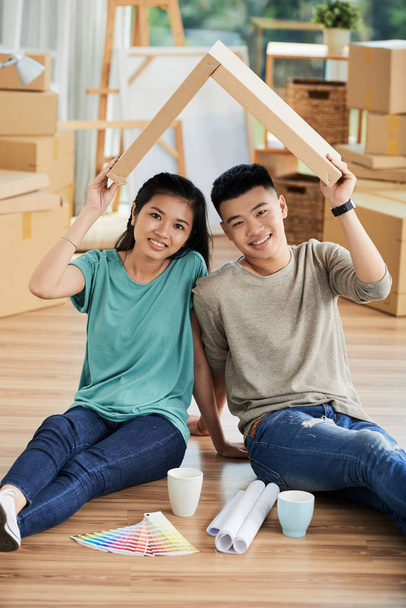 Portrait de jeune couple asiatique assis sur le sol tenant un panneau de bois sous la forme d'un toit au-dessus de leurs têtes et choisissant la couleur
 - Photo, image