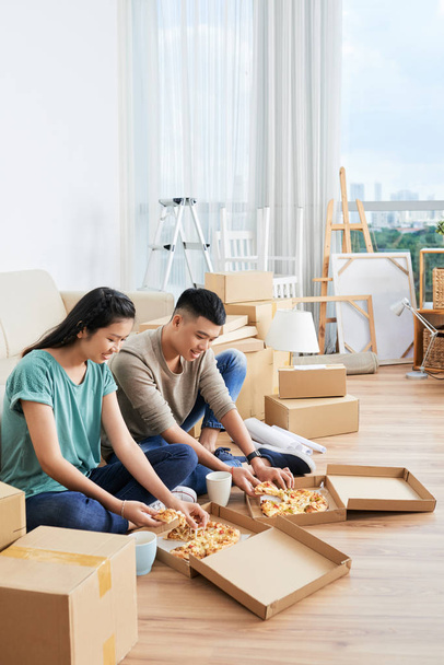 アジアの若いカップルは、彼らが新しい家に引っ越した後、一緒に座ってピザを食べる食べ物を取り除きます - 写真・画像