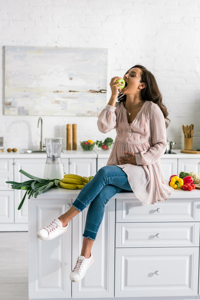 attractive pregnant woman sitting on table and eating apple  - Photo, image