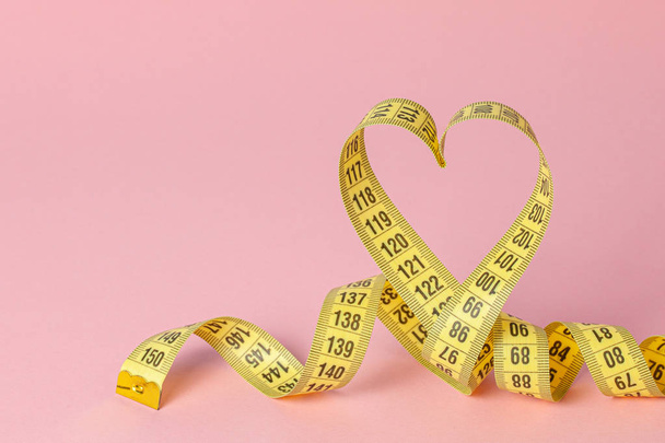 Gele meetlint in de vorm van een hartje op een roze achtergrond. Het concept van gewichtsverlies voor de normale werking van het hart en lichaam. Kopieer ruimte voor tekst. - Foto, afbeelding