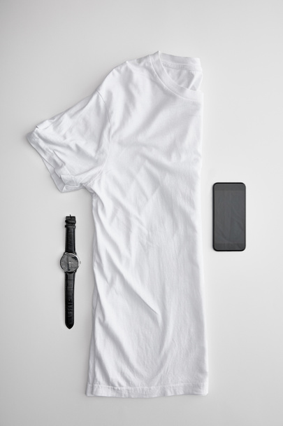 flache Lage mit in der Mitte gefaltetem Blanko-T-Shirt in der Nähe von Uhren und Smartphone auf weißem Hintergrund - Foto, Bild