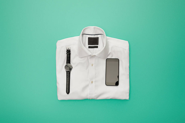 top view of watches and smartphone on plain white folded shirt on turquoise background - Foto, Imagen
