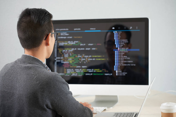 Вид спереду молодого програміста, який сидить за столом перед комп'ютерним монітором і розробляє нову програму в офісі
 - Фото, зображення
