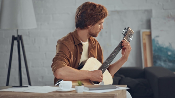 młody rude mężczyzna gra na gitarze akustycznej w salonie - Materiał filmowy, wideo