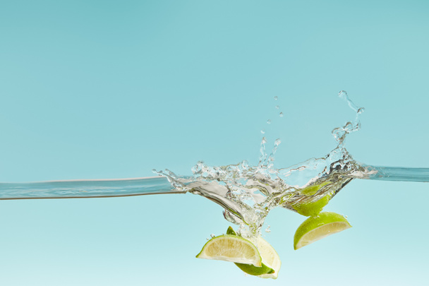 ripe lime pieces falling deep in water with splash on blue background - Photo, Image