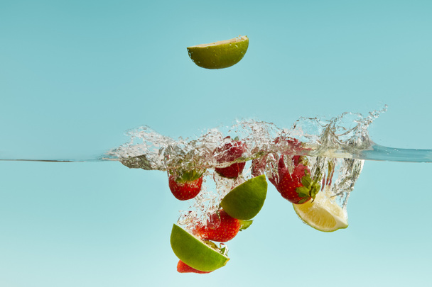 ripe lime pieces and strawberries falling deep in water with splash on blue background - Photo, Image