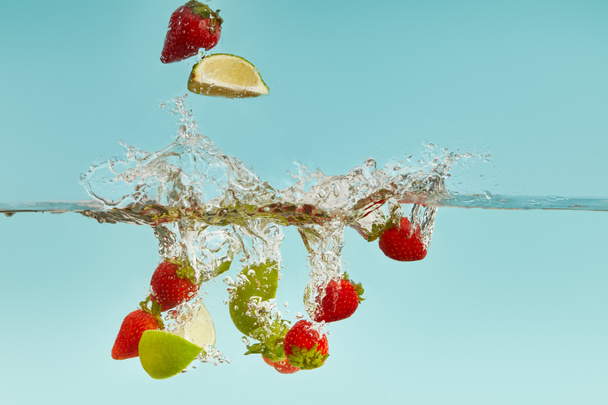 lime pieces and strawberries falling deep in water with splash on blue background - Photo, Image