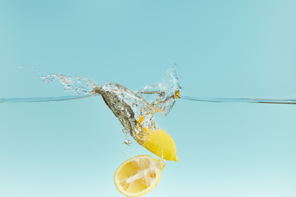 geschnittene Zitronen fallen tief ins Wasser mit Spritzer auf blauem Hintergrund - Foto, Bild
